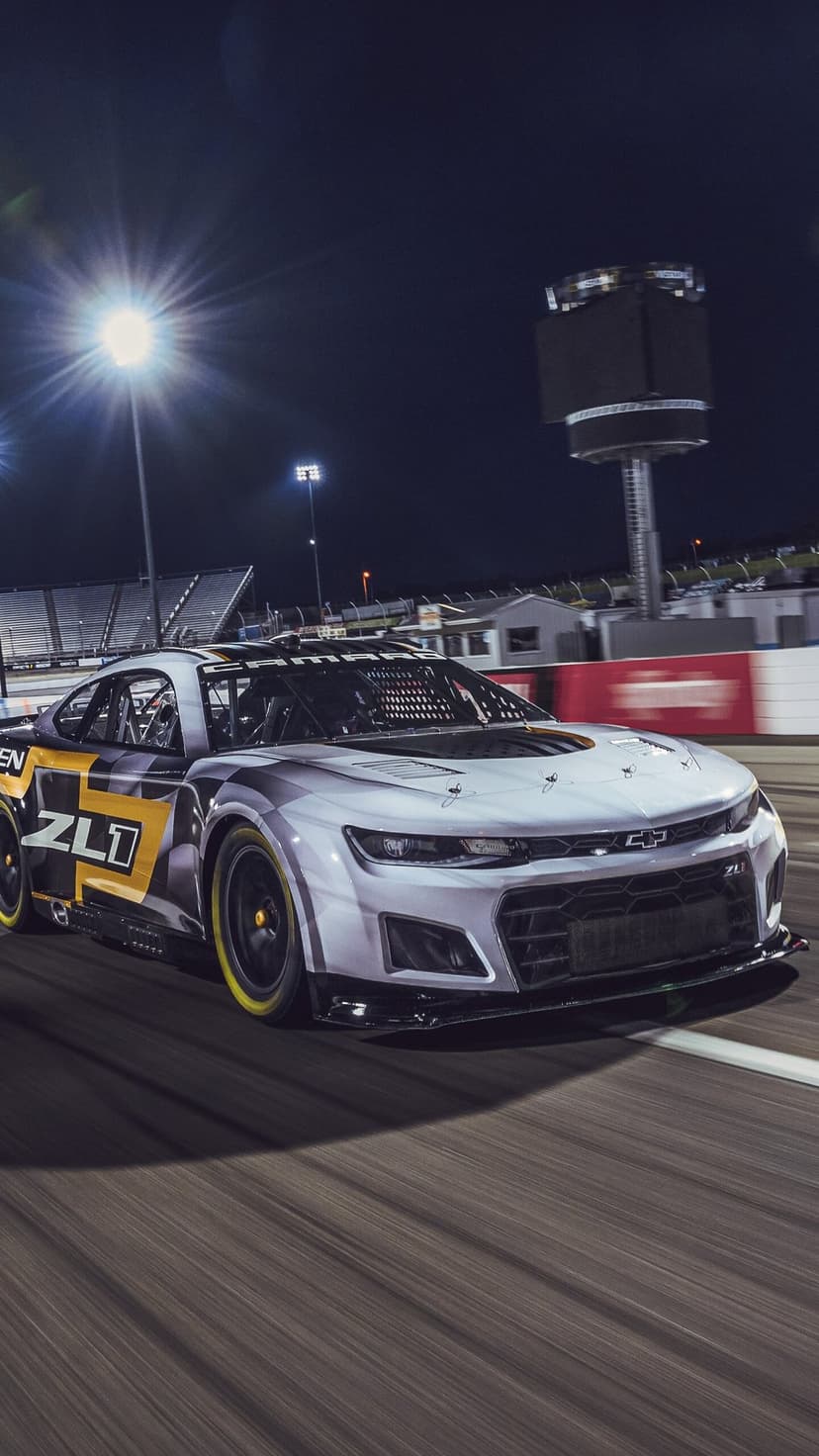 Silver Chevrolet Camaro ZL1 race car speeding on a track at night under bright stadium lights.