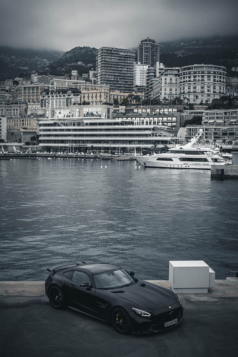 Black sports car parked on a stone pier facing luxury yachts and city architecture under a moody overcast sky.