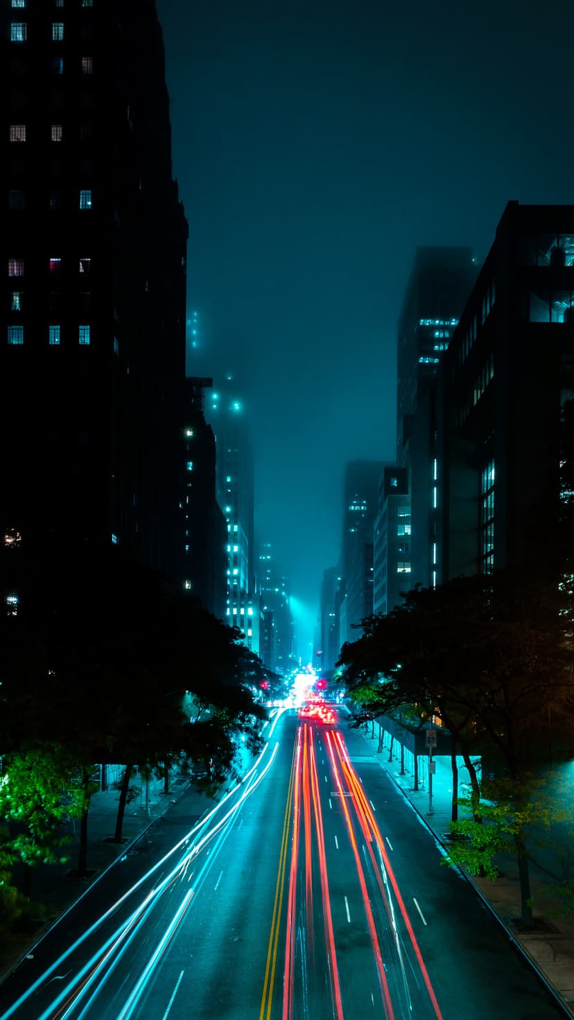 Radiant red light trails flow down a misty urban street at night, framed by towering dark skyscrapers.