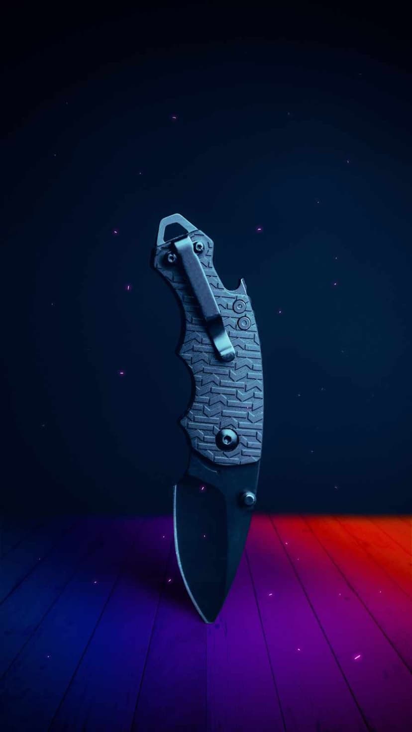 Tactical folding knife with a black serrated blade and textured gray handle on wood with red and blue light.