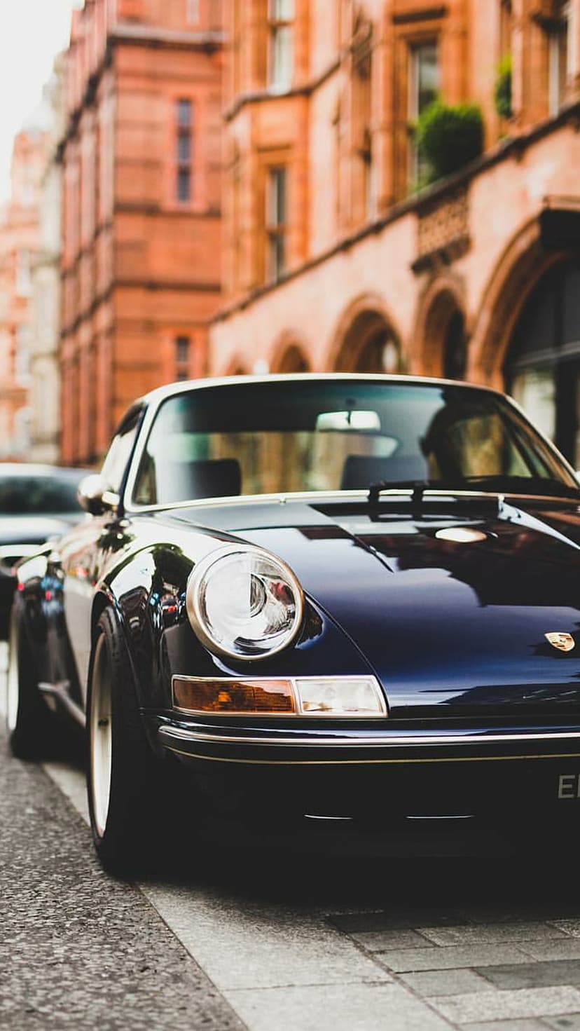 Dark blue vintage Porsche parked on grey cobblestones in front of a red brick building with arched windows.