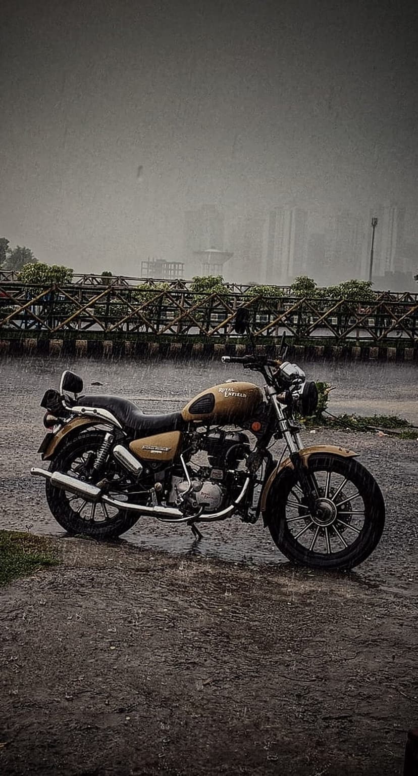 Royal Enfield motorcycle parked on a wet urban road during heavy rain with a wooden bridge in the background.