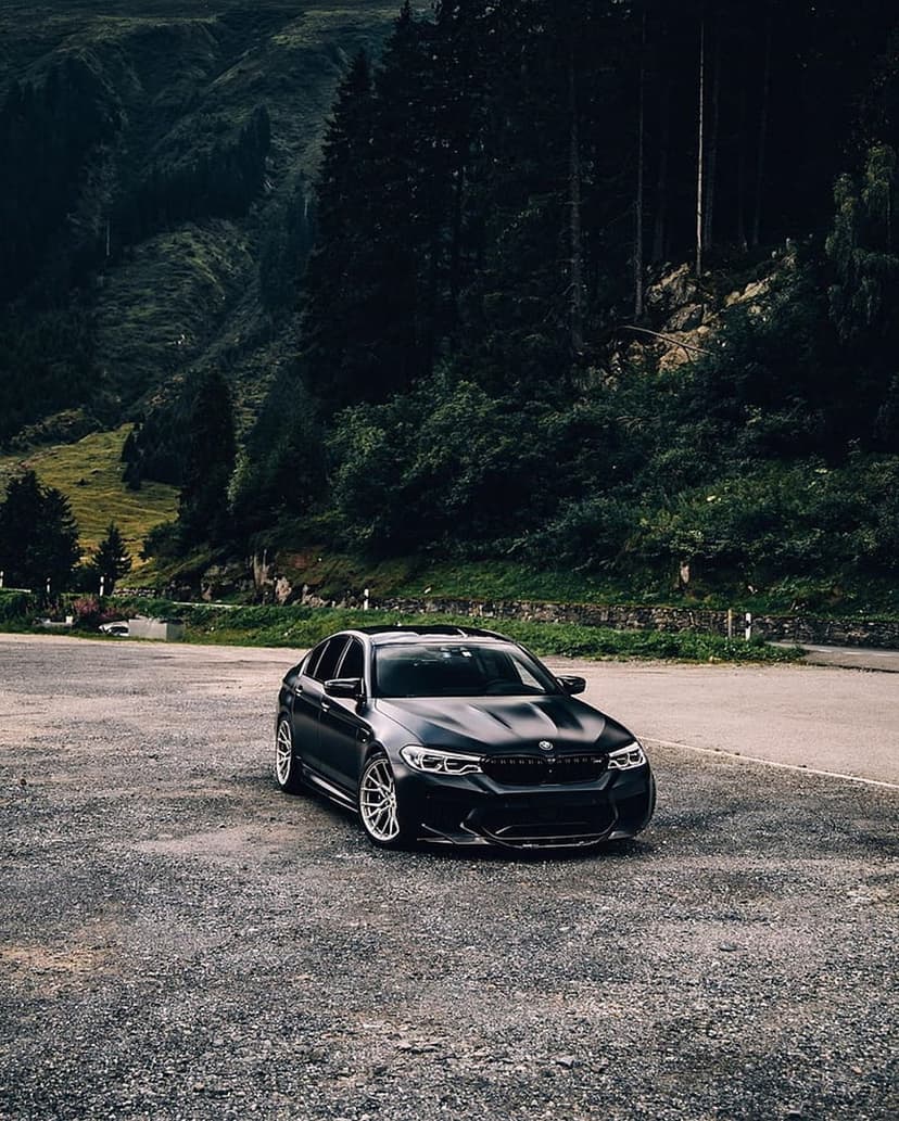 Black BMW M5 sedan parked on a rocky mountain road against a backdrop of steep green evergreen forests.