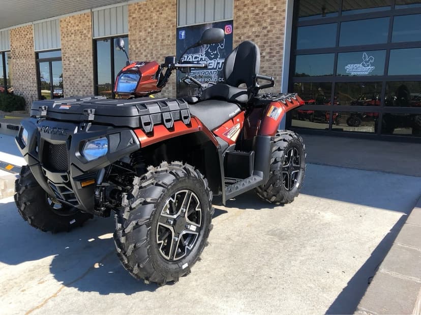Metallic orange Polaris ATV with aggressive mud tires parked on concrete in front of a brick building.