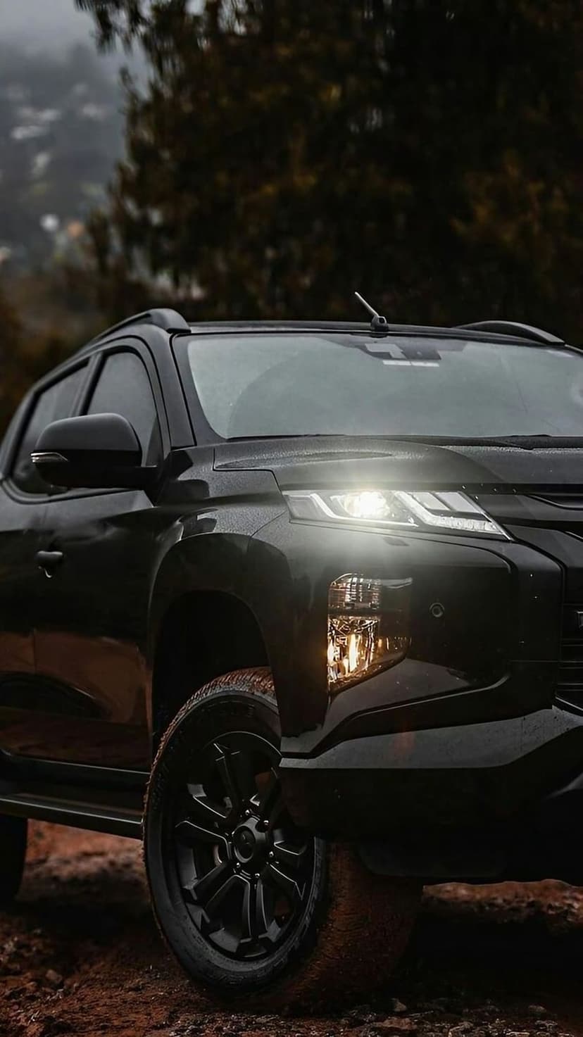 Black Mitsubishi Triton pickup truck with glowing headlights driving across a dusty off-road trail at dusk.