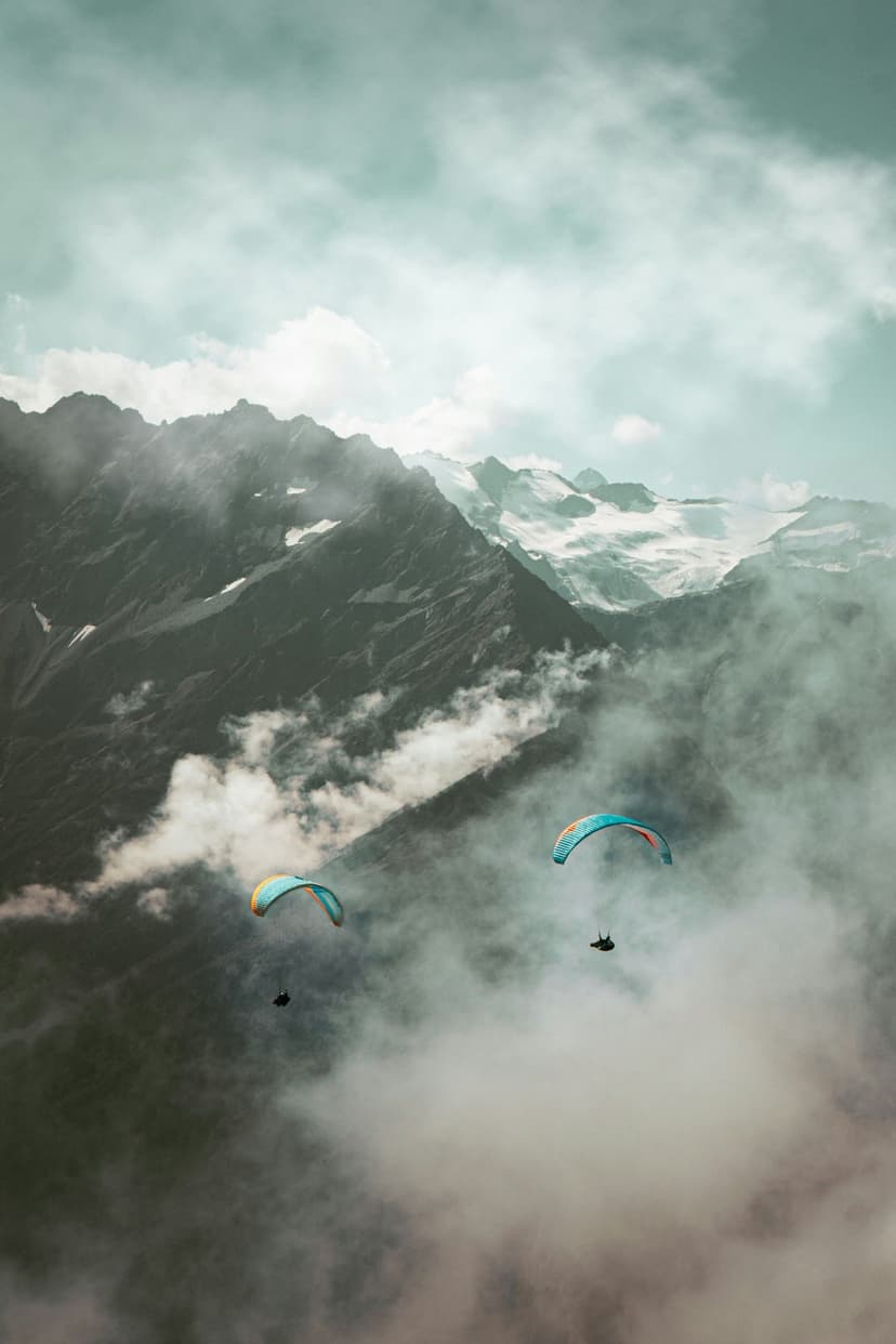 Paragliders soar through dense mountain fog above sharp snowy ridges and deep alpine valleys during a hazy day.