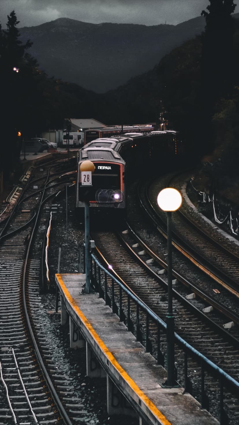 Locomotive headlamp glows on a curving track through a dark forest with foggy mountain peaks visible at twilight.