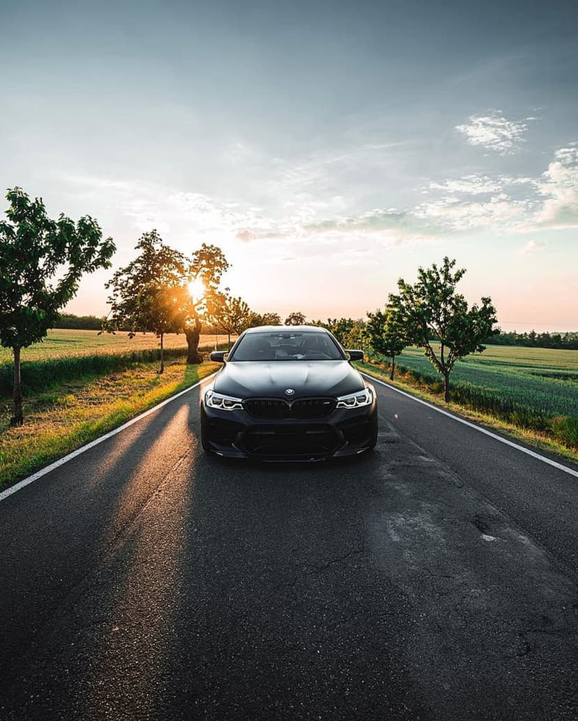 Black BMW M5 sedan drives on a rural road during a golden hour sunset with trees casting long shadows.