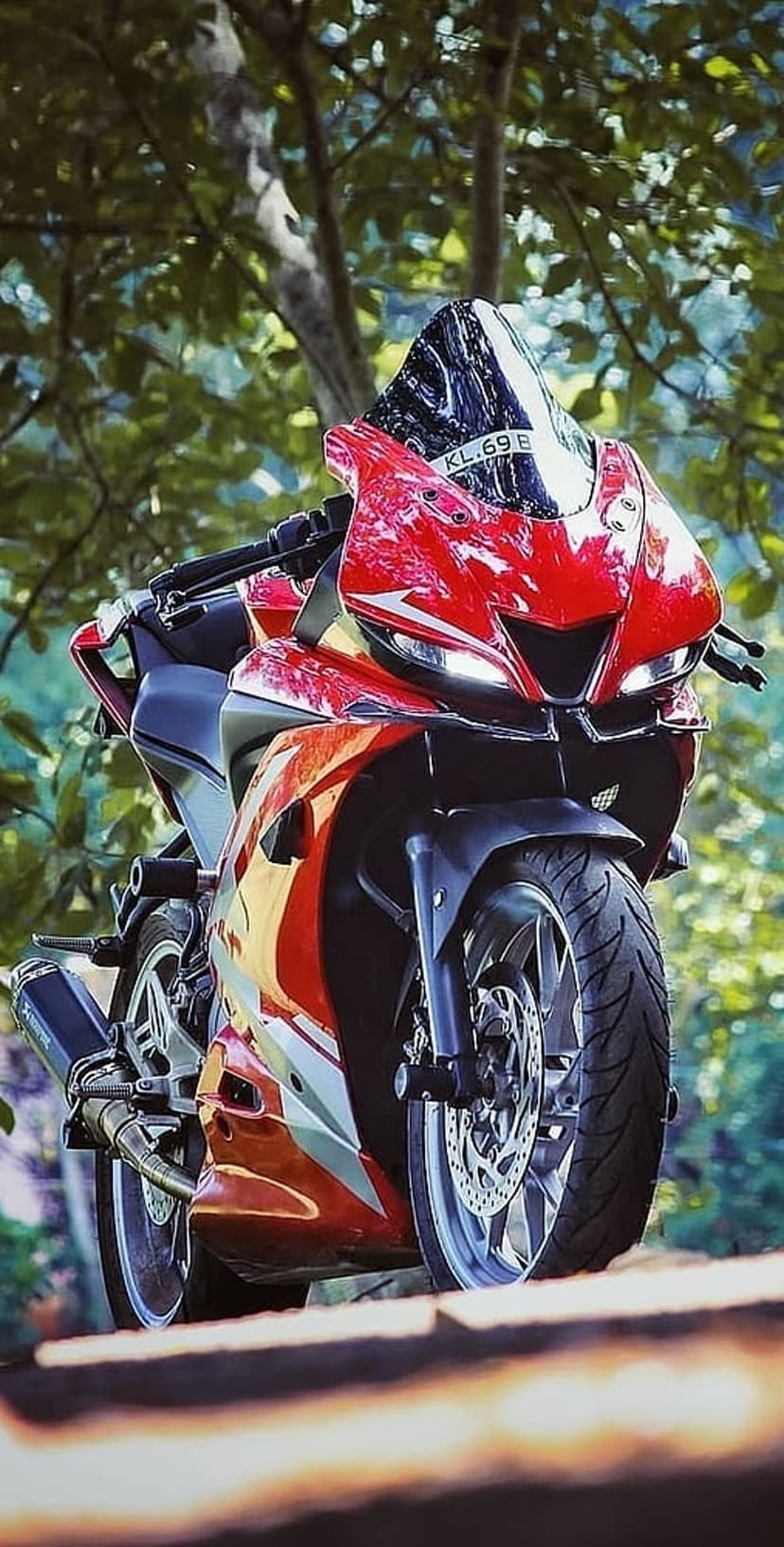 Red Yamaha R15 sportbike with a clear windshield parked outdoors against a soft green leafy background.