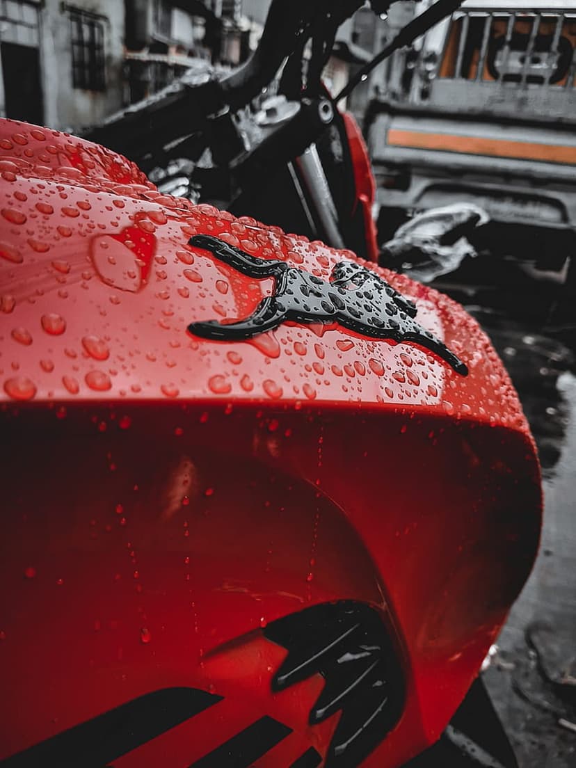 Red motorcycle fuel tank covered in water droplets with a black jaguar emblem set against a blurred city street.