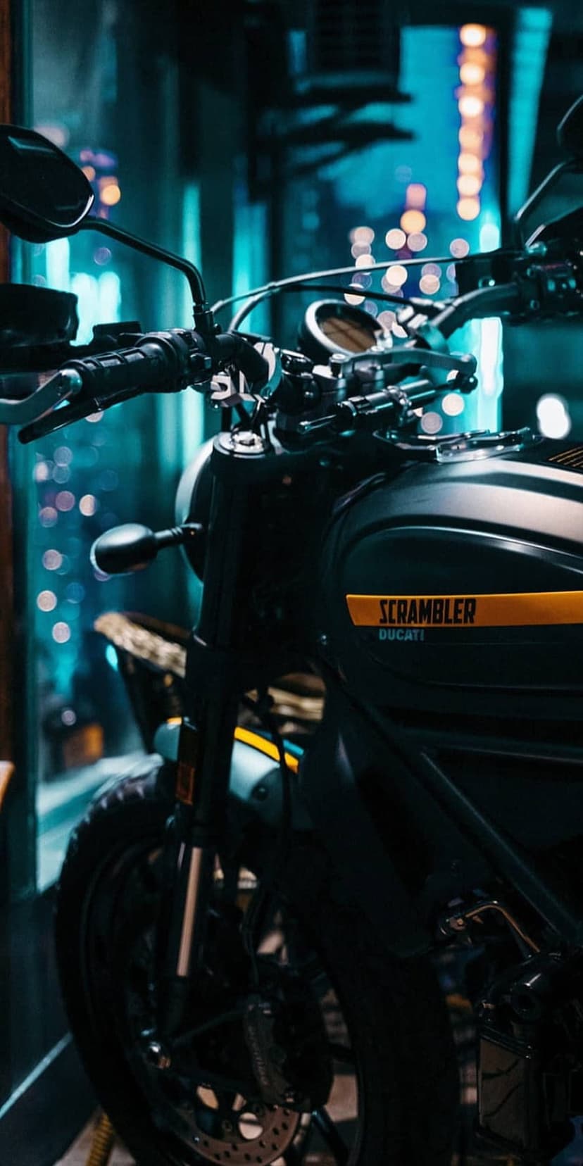 Black Ducati Scrambler motorcycle parked in a city at night with colorful bokeh lights blurred in the background.