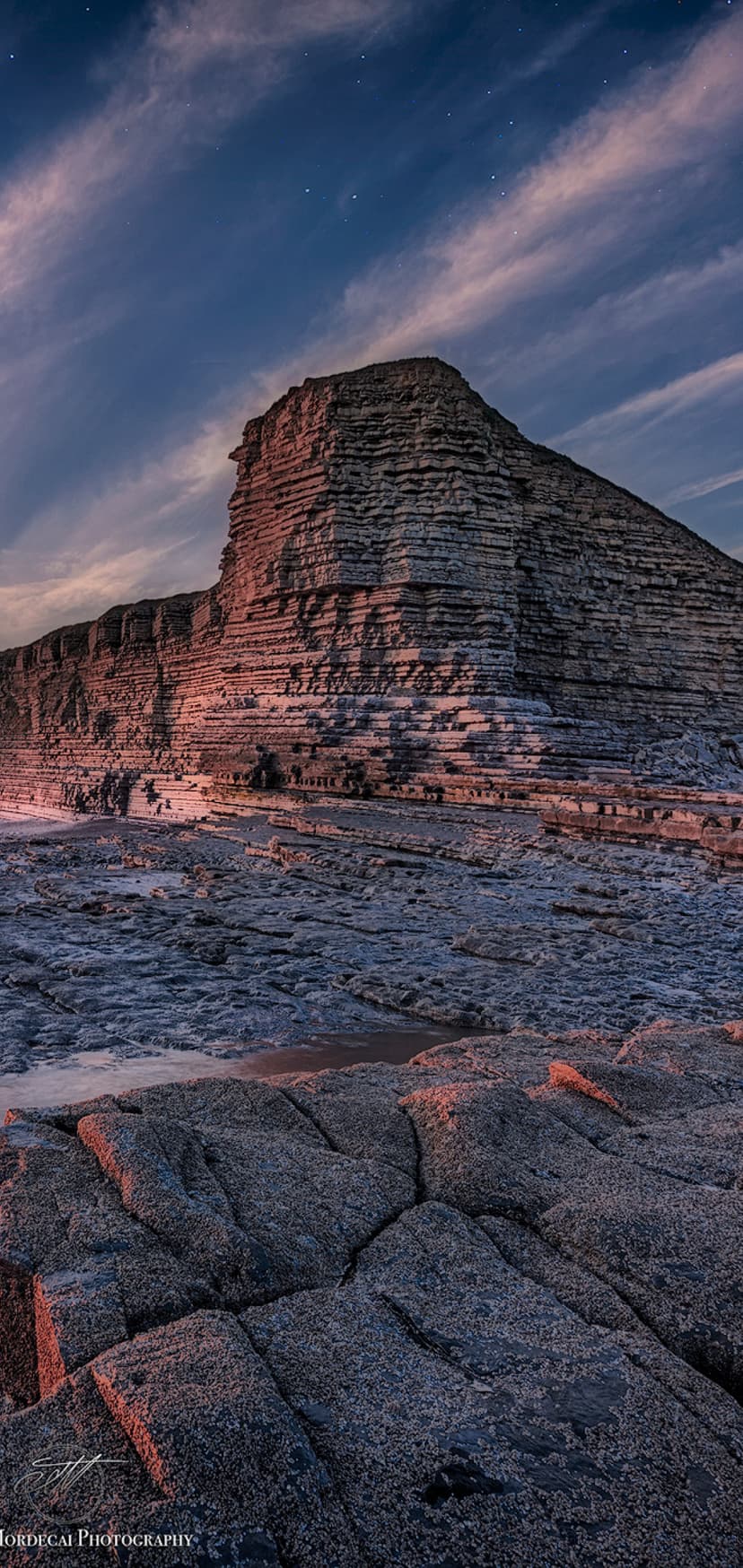 Stratified rock cliff rises toward a starry twilight sky with orange horizon glows above a rugged coast.