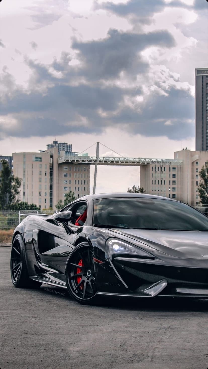Black McLaren 600LT supercar parked on asphalt with a modern suspension bridge and city skyline background.