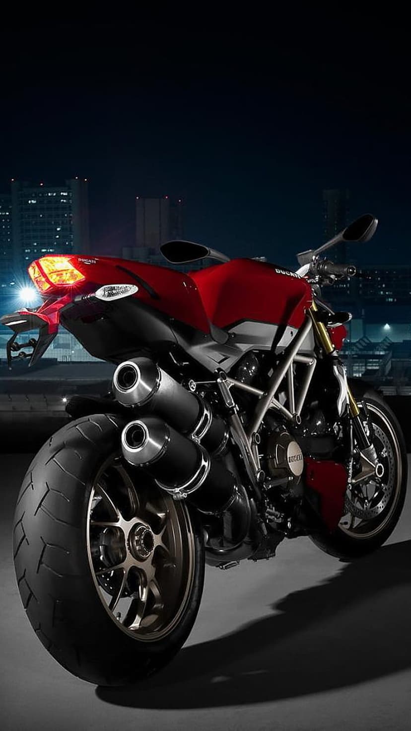 Red Ducati motorcycle rear wheel and dual exhausts against a blurred night city skyline with glowing lights.