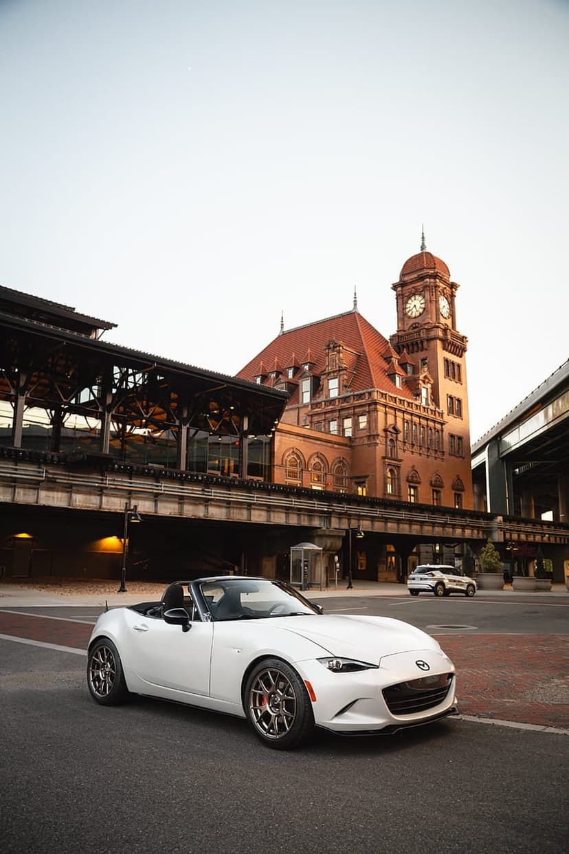 White Mazda Miata convertible parked on a city street in front of a historic stone train station clock tower.