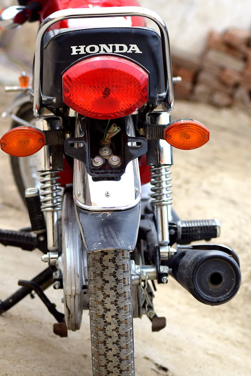 Honda motorcycle rear view with a red taillight, chrome details, and orange signals on a gravel surface.