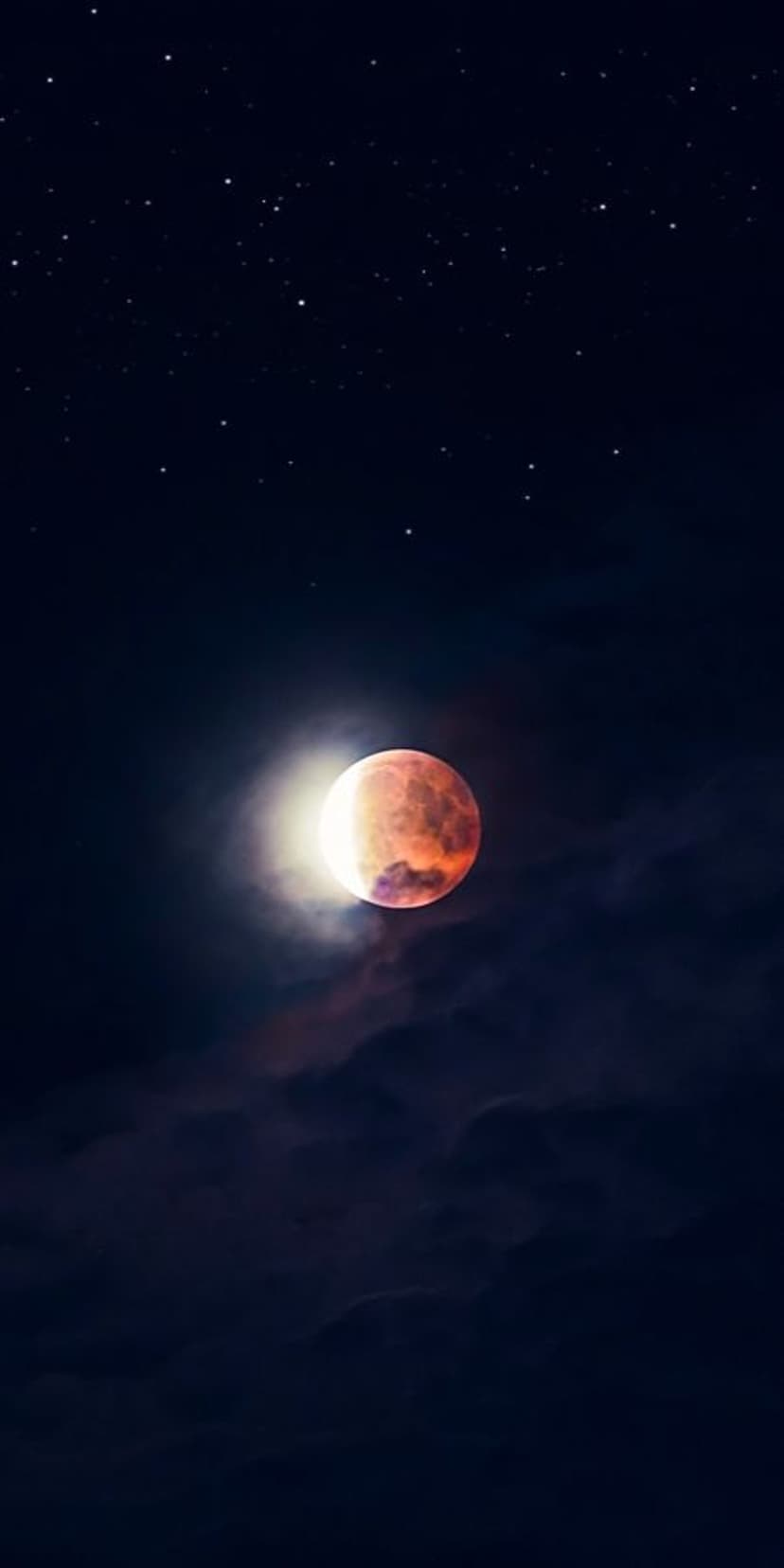 Blood moon glowing in a deep indigo sky with wispy clouds and stars during a partial lunar eclipse transition.
