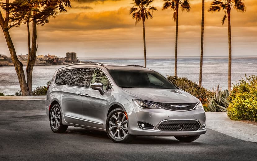 Silver Chrysler Pacifica parked on a coastal road overlooking the ocean under an orange sunset sky with palm trees.