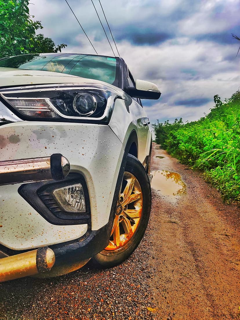 White car with golden rims parked on a muddy gravel road surrounded by dense green bushes under a cloudy sky.