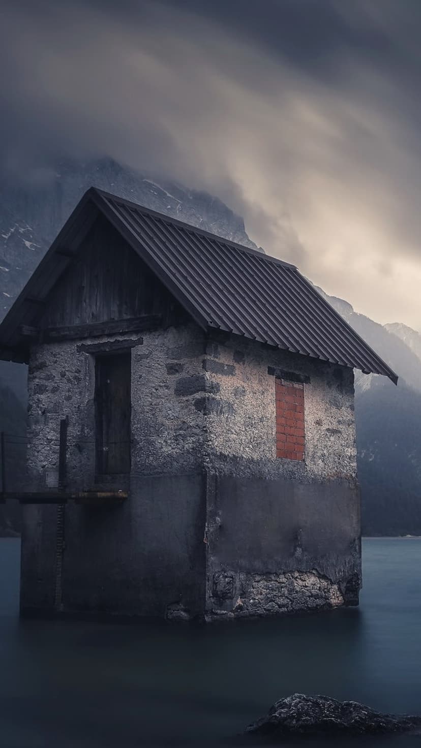 Stone boathouse on a still dark lake with foggy mountains rising behind it under a heavy overcast gray sky.