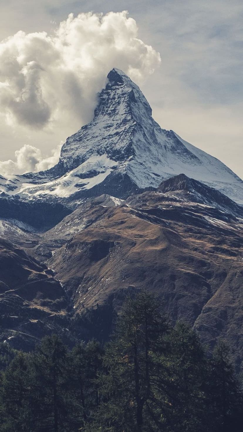 Matterhorn peak with snow-covered ridges rises through swirling clouds above dark green forest slopes.