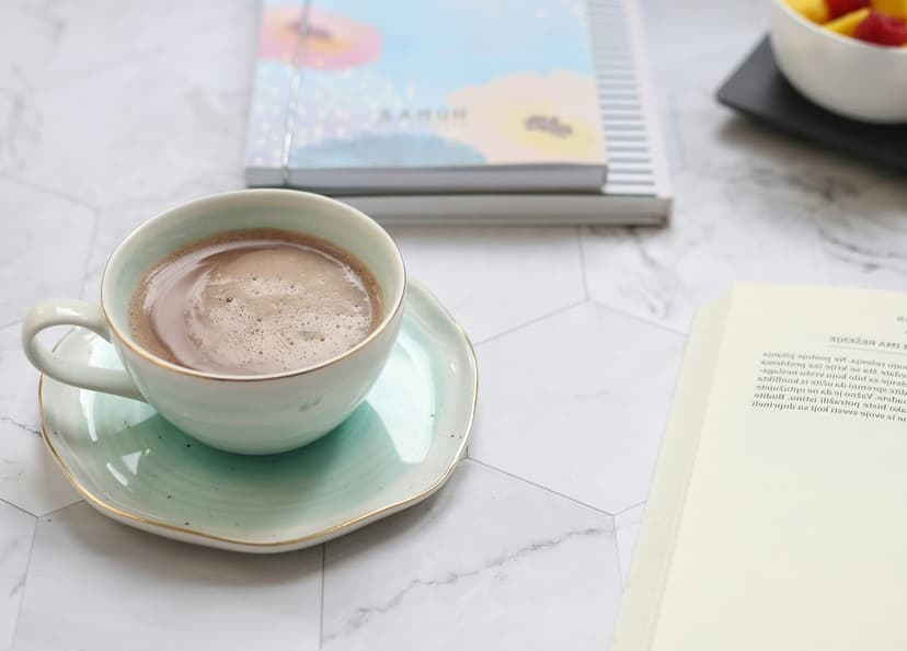 Hot chocolate mug on a mint green saucer next to an open book and journals on hexagonal marble tiles.