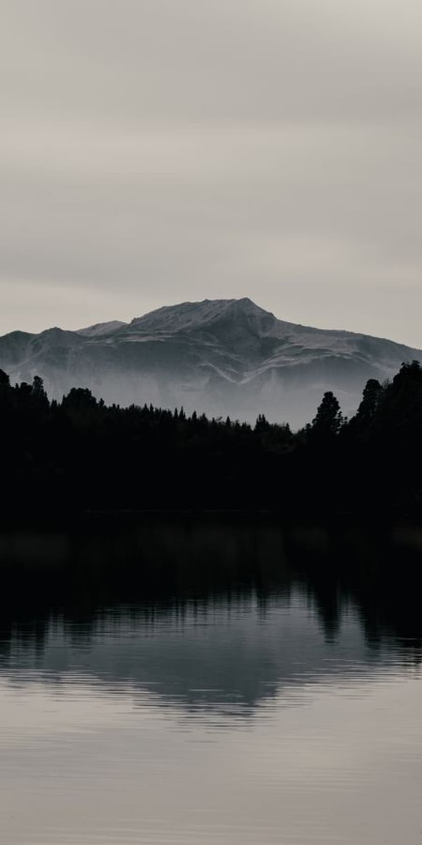 Dark mountain silhouettes and a dense forest reflect in the mirror-like surface of a misty lake during twilight.