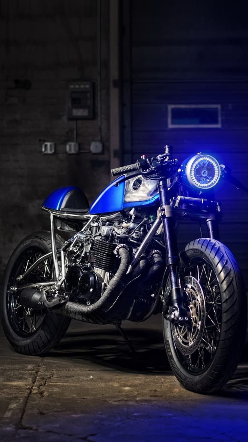 Custom blue cafe racer motorcycle with a glowing circular LED headlight parked on a concrete garage floor at night.