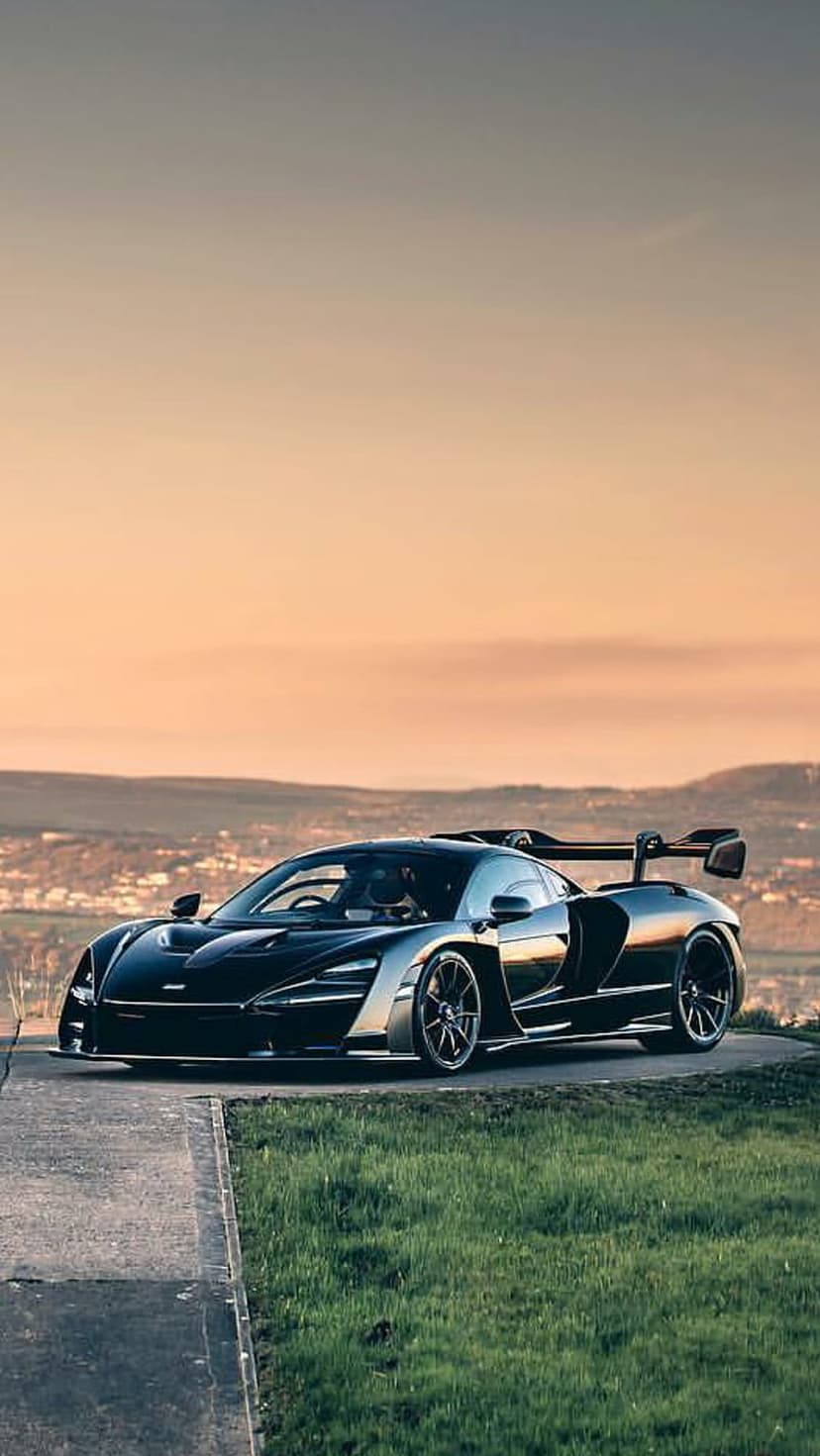 Black McLaren Senna supercar with a large rear wing parked on a hilltop road overlooking a valley during sunset.