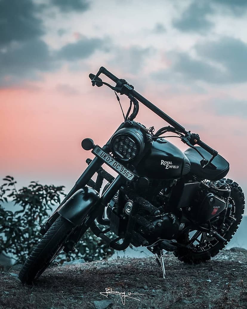 Black Royal Enfield motorcycle with a glowing LED headlight parked on rocks under a soft pink sunset sky.