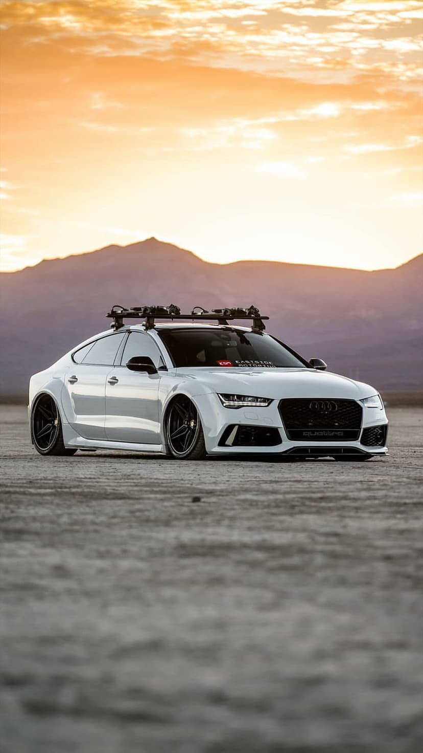 White Audi RS7 with roof racks parked on a flat desert plain under a vibrant orange and purple sunset sky.
