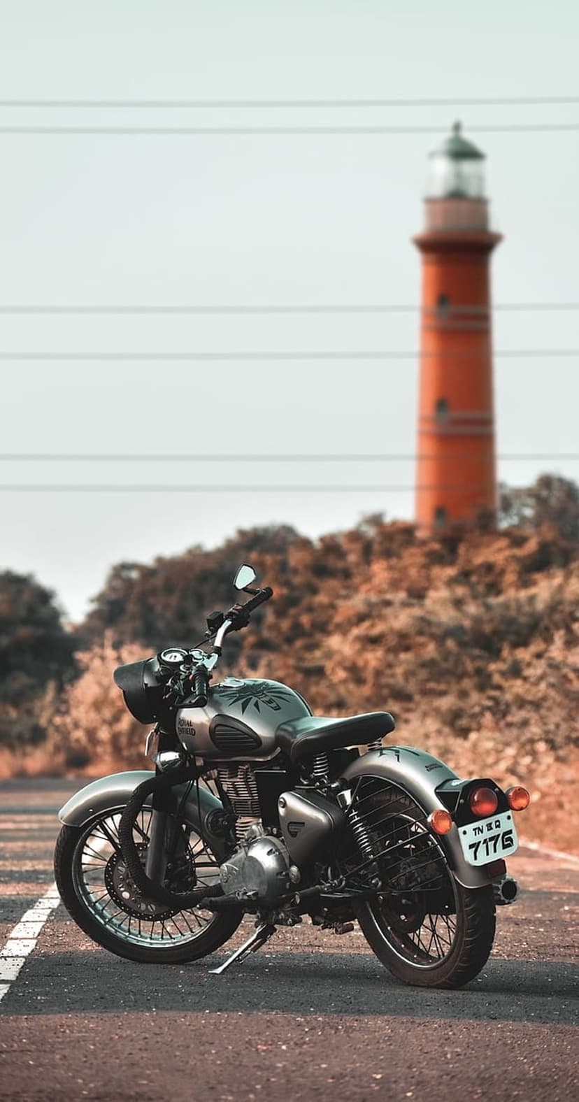 Silver Royal Enfield motorcycle parked on an asphalt road with a white lighthouse and red foliage background.