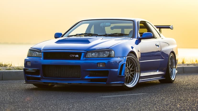Blue Nissan Skyline GT-R R34 parked on a road at sunset with golden light and BBS wheels.