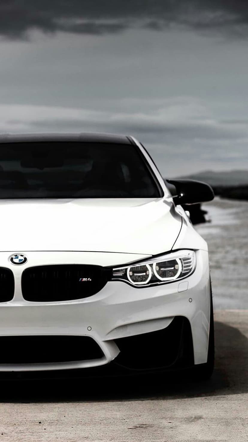 White BMW M4 coupe with an aggressive front grille parked on a coastal road under a dark, stormy sky.