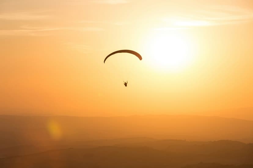 Paraglider silhouette soaring over rolling hills against a deep orange sunset sky with a warm golden glow.