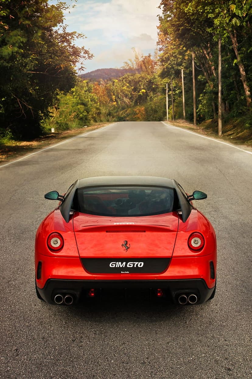 Red Ferrari 599 GTO rear view parked on an asphalt road with hazy green mountains and trees in the distance.