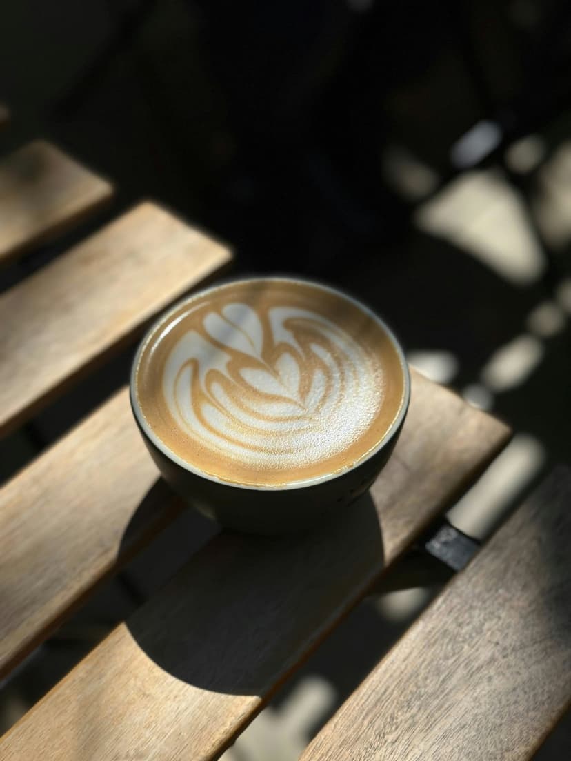 Rosetta latte art in a ceramic cup sits on a rustic wooden table with bright sunlight and dappled shadows.