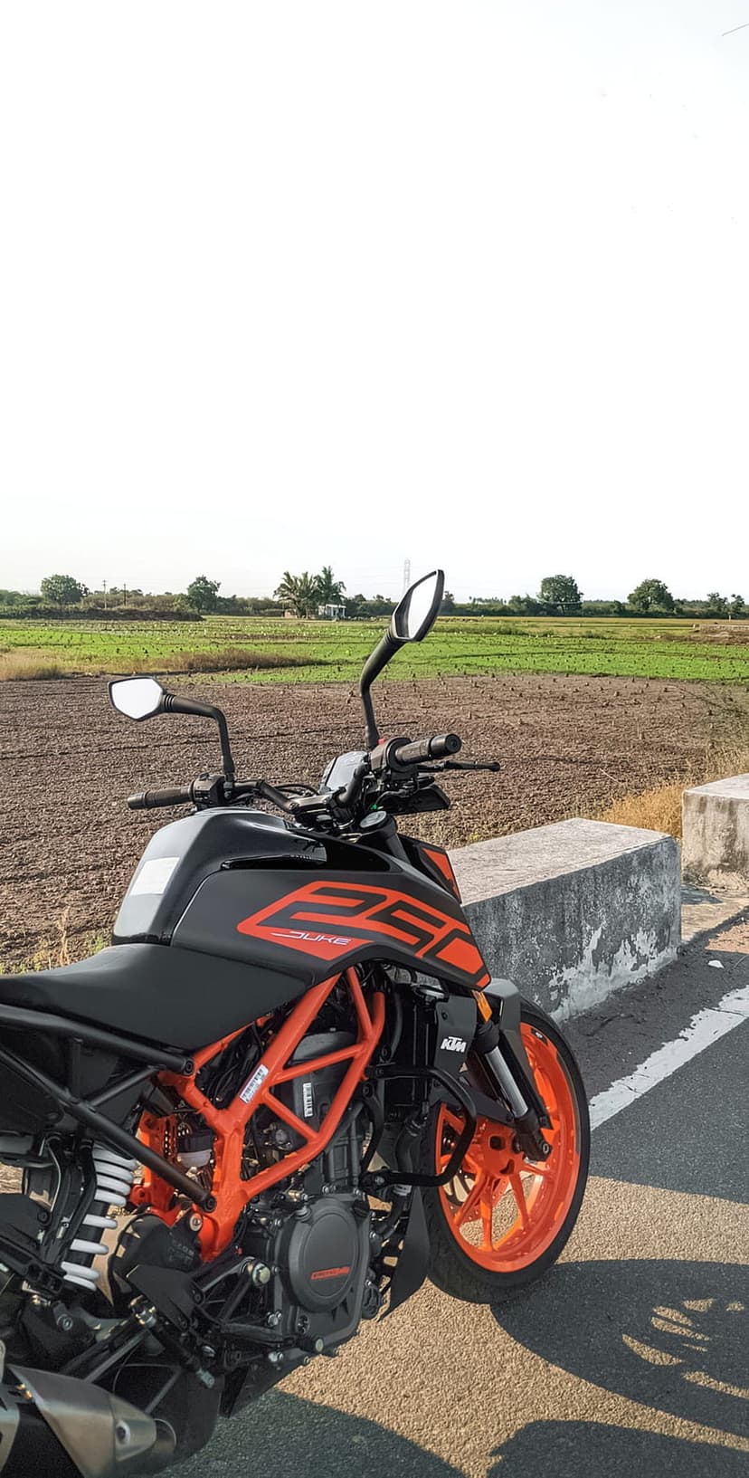 KTM 250 Duke motorcycle with an orange frame parked on a rural roadside next to green agricultural fields.