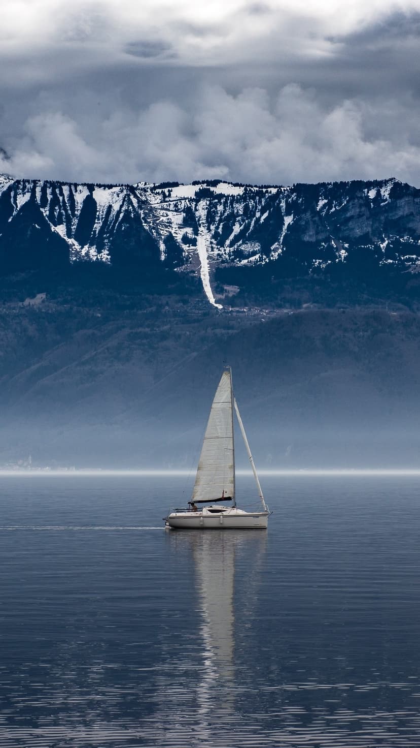 White sailboat drifts on a glassy mountain lake under snow-capped peaks and overcast skies in a vertical view.