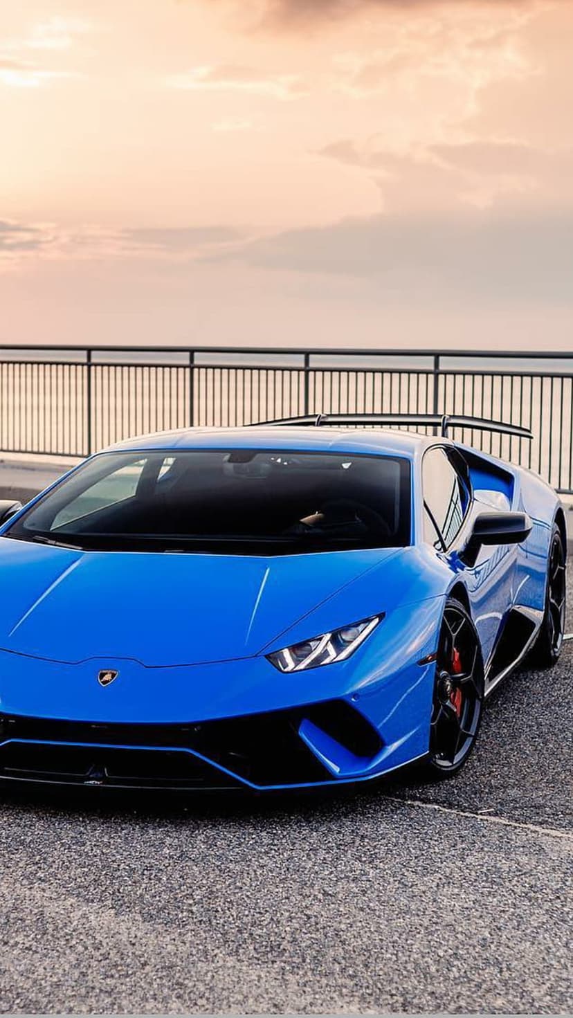 Blue Lamborghini Huracan parked on asphalt at sunset with a rear wing and soft pastel clouds in the background.