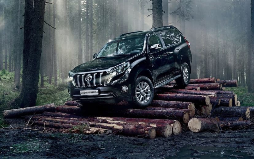 Black Toyota Land Cruiser Prado SUV parked on a pile of logs in a misty pine forest with sunlight rays.