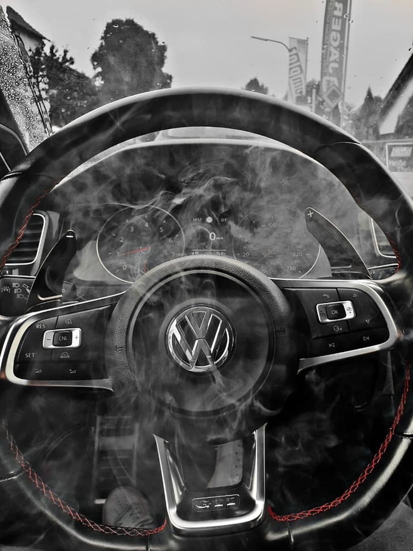 Volkswagen leather steering wheel with red stitching surrounded by smoke in front of a digital car dashboard.