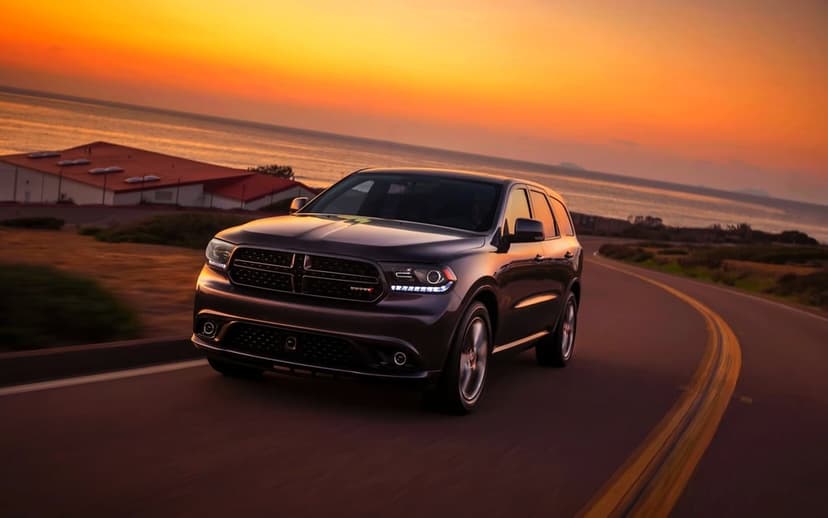 Dark metallic SUV driving on a winding coastal road during a vibrant orange sunset with calm ocean views.