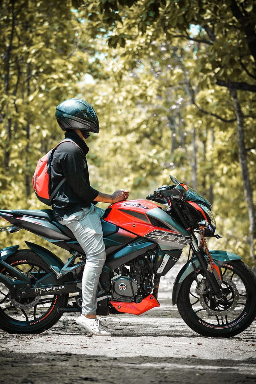 Biker in a black helmet and red backpack rides an orange motorcycle through a lush green forest on a dirt path.