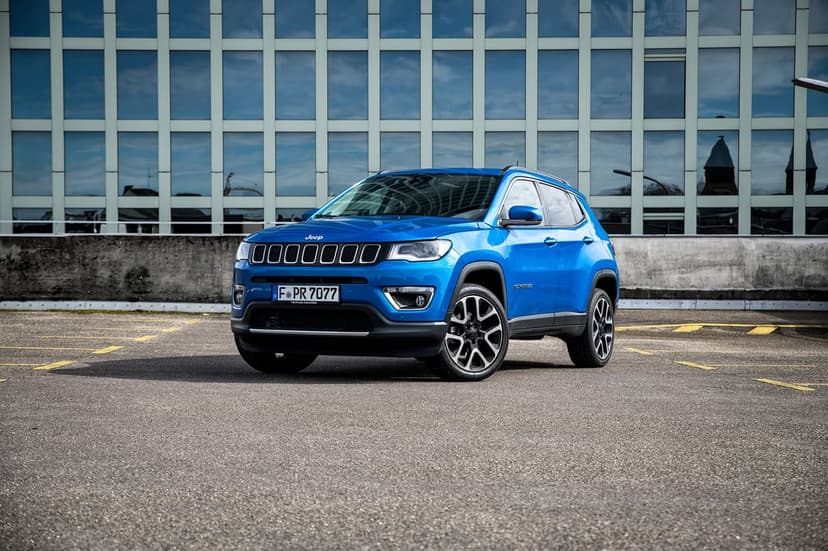 Blue Jeep Compass SUV parked on asphalt in front of a modern glass building with a grid-like window pattern.