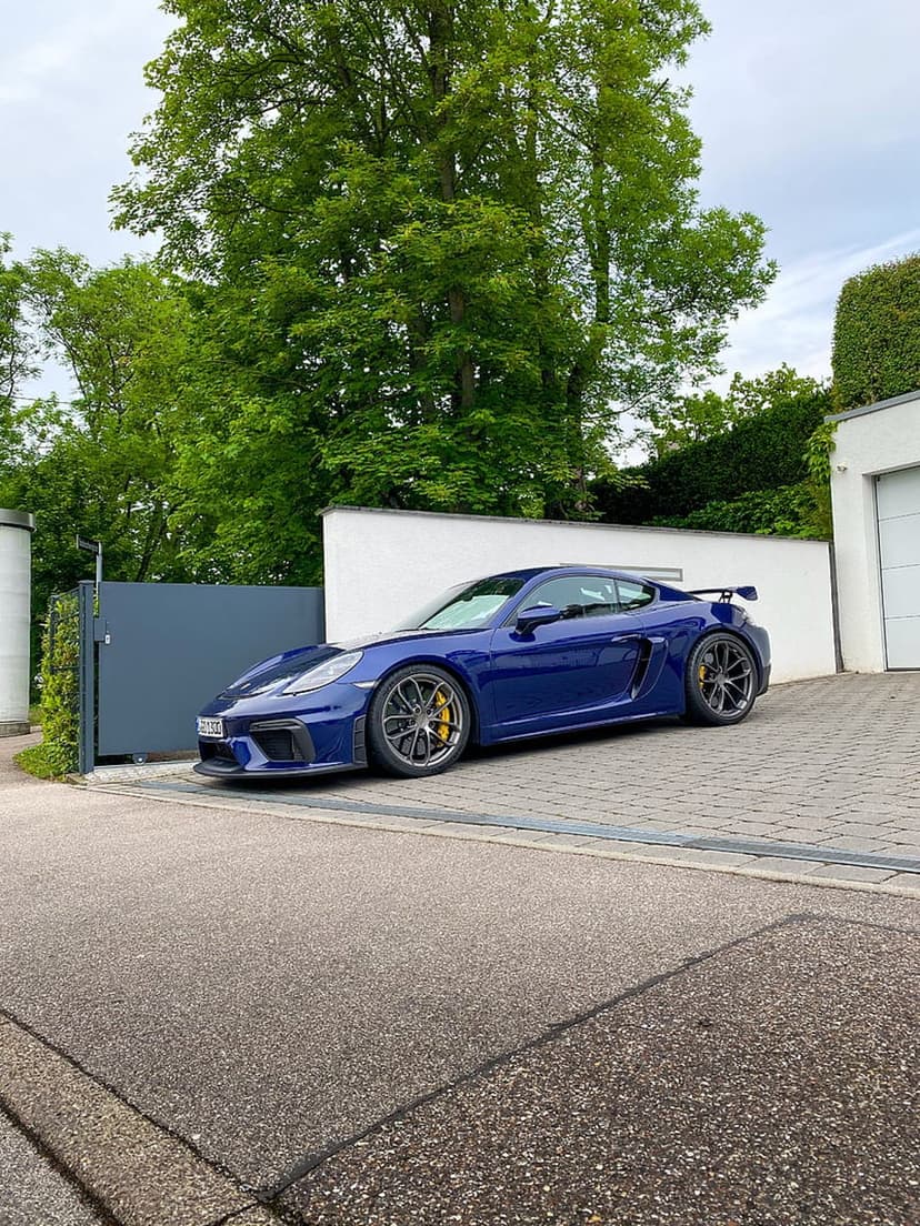 Blue Porsche Cayman GT4 parked on a stone driveway next to a minimalist white house with surrounding green trees.