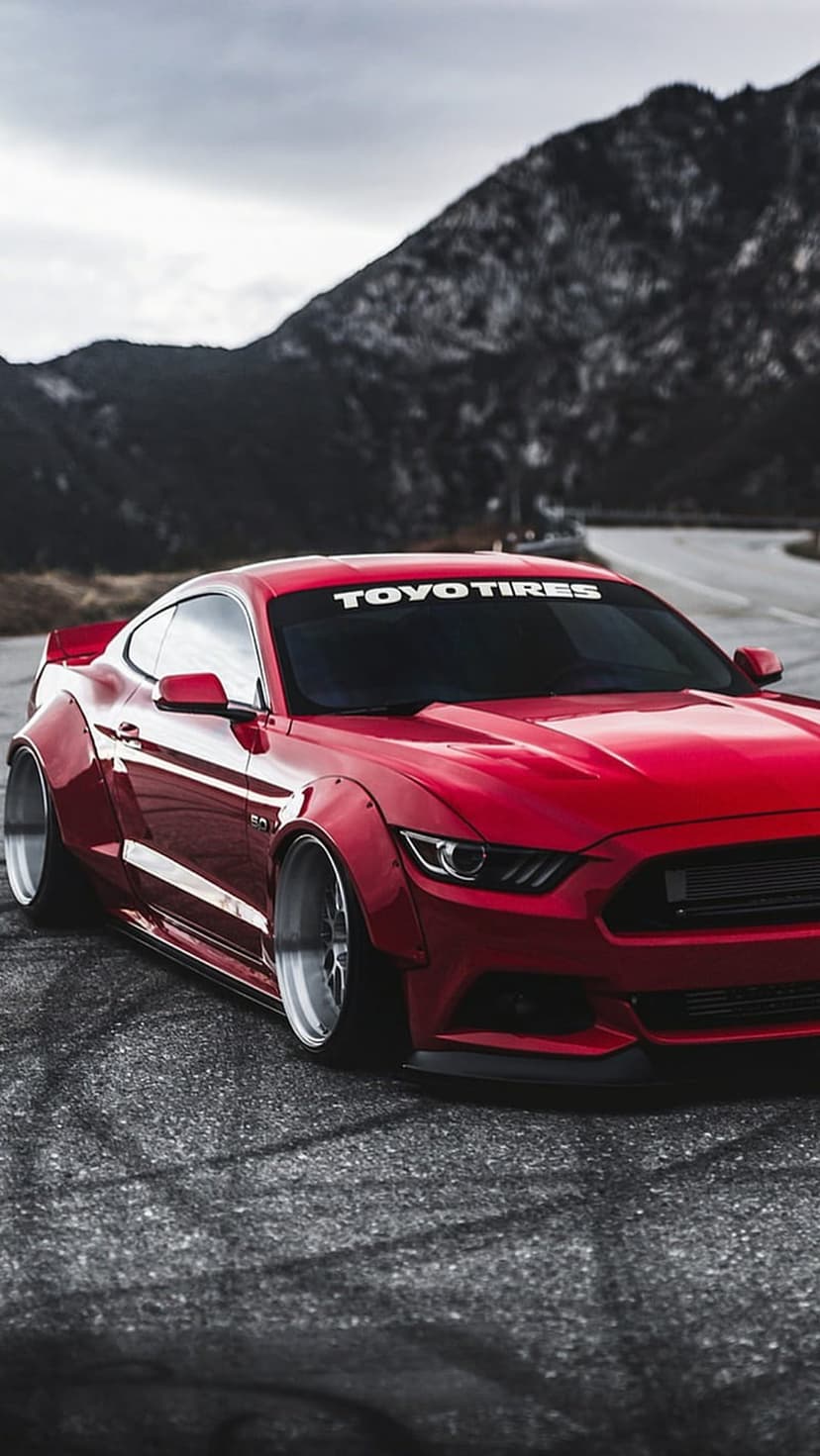 Red Ford Mustang with widebody kit and chrome wheels parked on a street with mountain peaks in the background.