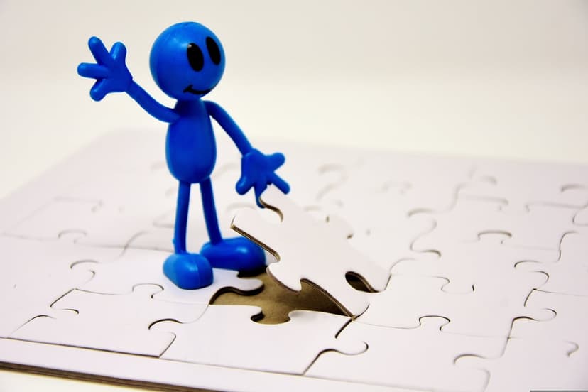Blue toy figure waves while placing the final piece into a white jigsaw puzzle on a bright surface.