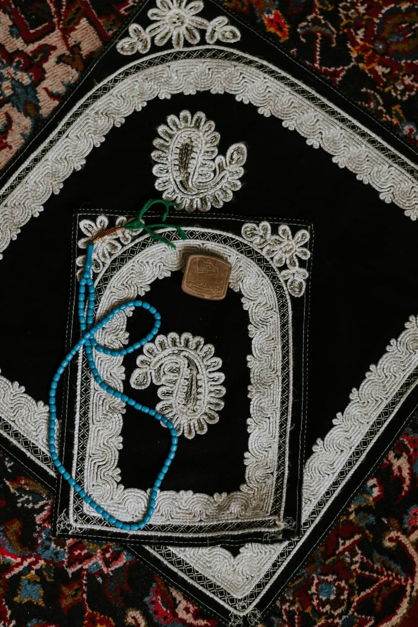 Black embroidered prayer mat with blue beads and a small brown Quran resting on a patterned Persian rug.