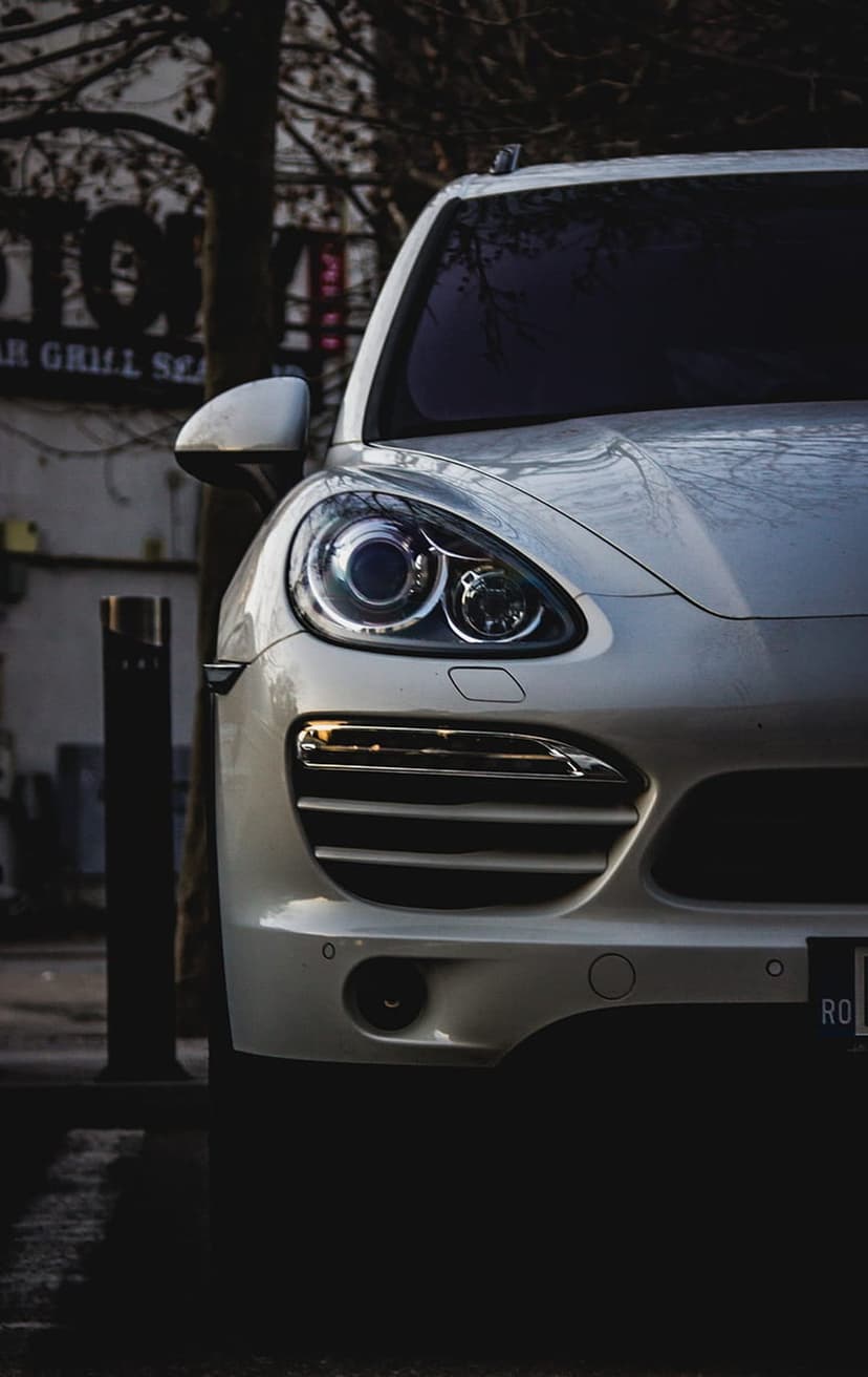 White Porsche Cayenne SUV front view showing LED headlights and chrome grille parked on a sunlit city street.